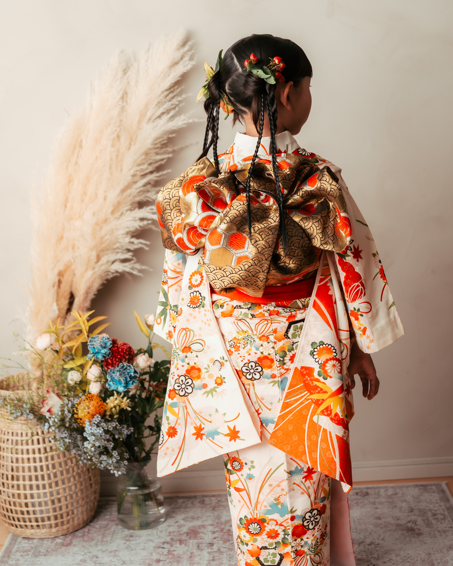 七五三用の華やかな白地の着物を着た7歳女の子の後ろ姿。金の帯と編み込みヘアスタイルが特徴的