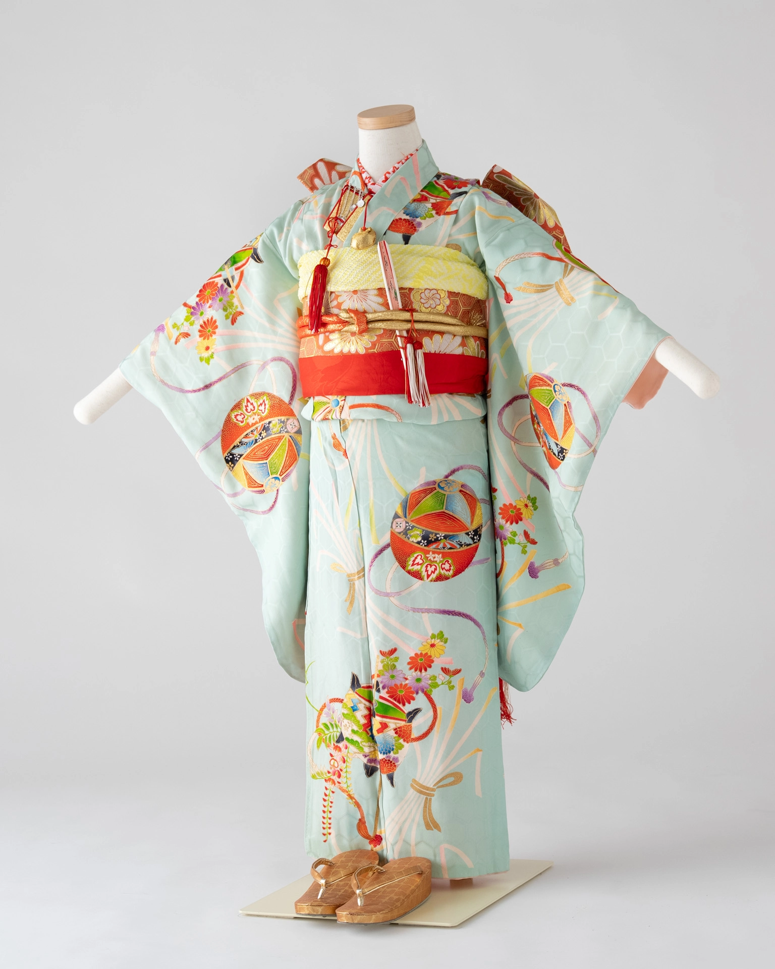 ミントグリーンの地に色とりどりの鞠と花柄が描かれた7歳女児用の七五三着物。赤い帯締めと金色の帯が特徴。
