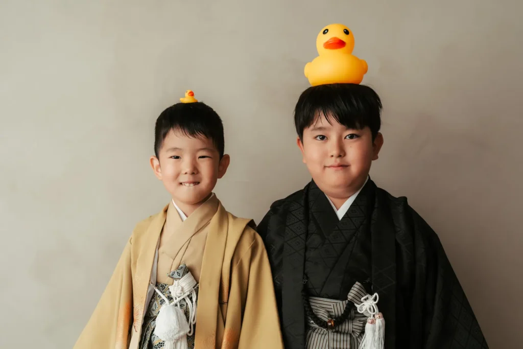 吉祥寺の家族の風景フォトスタジオで撮影した男の子2人の着物姿。ベージュと黒の羽織袴を着た兄弟が、大小の黄色いアヒルを頭に乗せて並ぶ、遊び心あふれる記念写真。